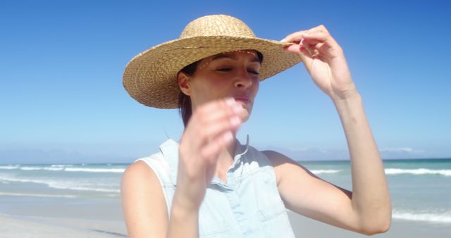 Young woman enjoying a sunny day at the beach is adjusting her straw hat. The clear blue sky and ocean waves create a relaxing, peaceful atmosphere. This image is perfect for travel brochures, vacation package websites, summer fashion promotions, and lifestyle blogs focusing on beach activities and relaxation.