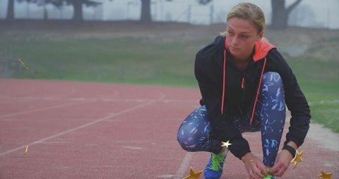 Squatting young female athlete tying neon laces on running shoes on misty outdoor track