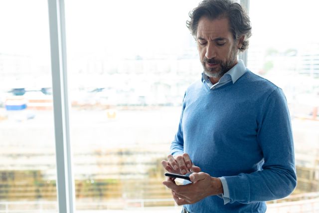 front view of mature Caucasian businessman using mobile phone near window in a modern office