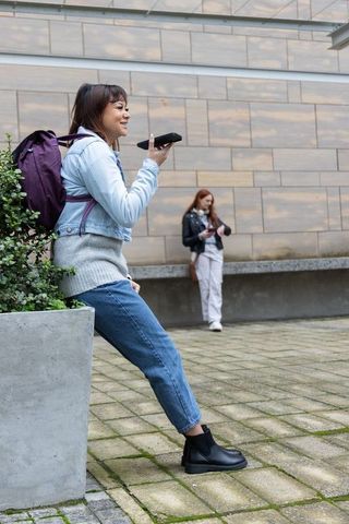 Young woman leaning on planter talking on smartphone while friend checking phone in courtyard