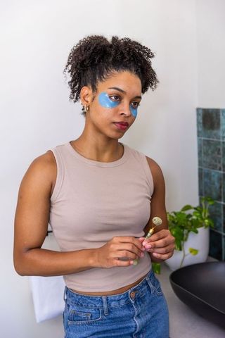 African american woman using under-eye patches and facial roller in bathroom