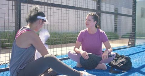 Two women sitting on blue padel court resting and chatting, wiping sweat, holding paddle
