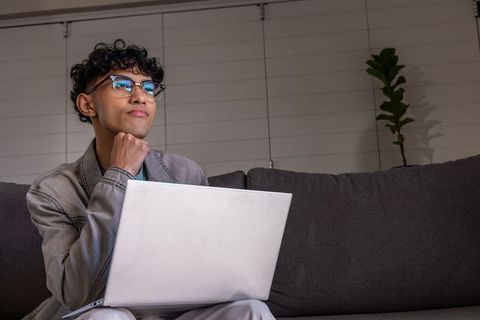 Thoughtful asian man working remotely on laptop while sitting on modern gray sofa