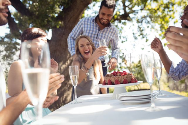 Group of friends celebrating a woman's birthday outdoors with cake and champagne. Everyone is laughing and enjoying the sunny day. Ideal for use in advertisements, social media posts, and articles about celebrations, friendship, and outdoor gatherings.