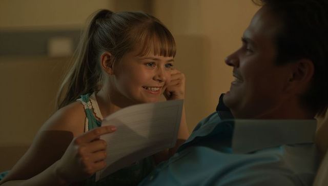 Smiling daughter leaning toward father on couch holding lined paper in cozy evening light