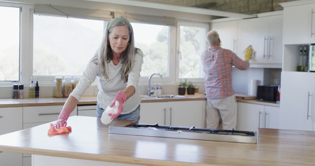Senior couple cleaning their modern kitchen at home, with woman wiping the kitchen counter and man cleaning cabinets. Ideal for themes related to household chores, teamwork, senior lifestyle, and domestic life. Useful in advertisements for cleaning products, articles on maintaining a clean home, or promotional materials for senior living communities.