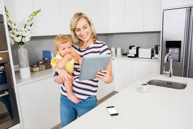 Mother holding her baby while using a digital tablet in a modern kitchen. The mother is smiling and casually dressed, highlighting a typical day in a household setting. The kitchen is spacious with white cabinets and contemporary appliances. This image can be used for parenting blogs, family-oriented advertisements, technology usage promotions, and articles on multitasking in daily life.