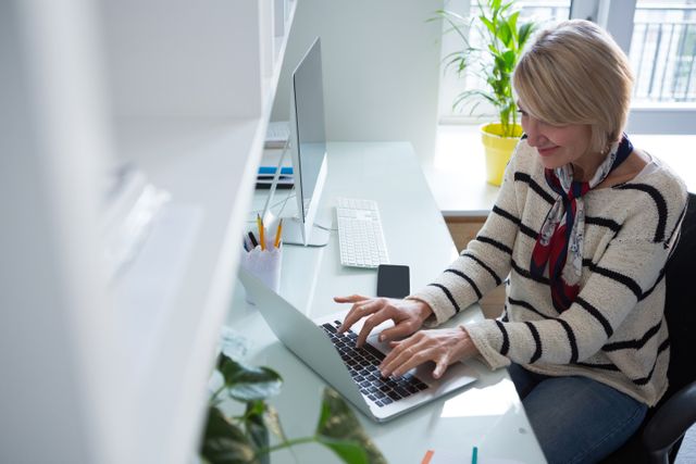 Woman sitting at desk in home office, typing on laptop, smiling. Modern workspace with plants and natural light. Ideal for illustrating remote work, productivity, home office setups, and professional women.