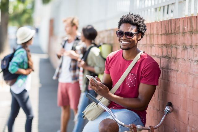 Happy young man sitting on bicycle using mobile phone with friends interacting background