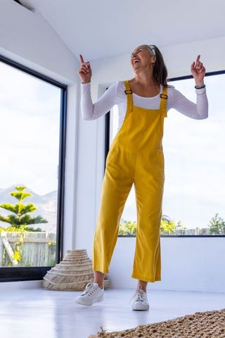 Mature woman dancing in yellow overalls in bright modern living room with large windows