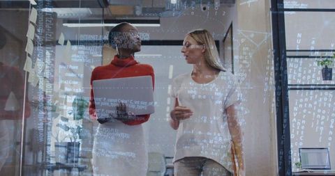 Two professionals walking and collaborating with laptop and tablet, code overlay on glass