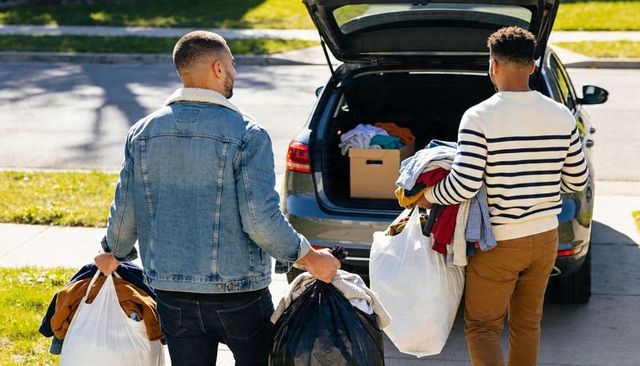 Two men donating clothes and loading hatchback trunk on sunlit suburban curbside