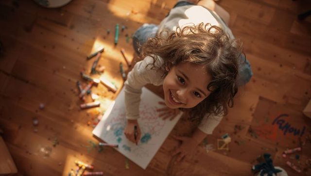 Curly-haired girl drawing with crayons on sunlit hardwood floor smiling upward joyful mood