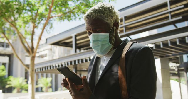 African american senior man wearing face mask using smartphone in corporate park. hygiene and social distancing during coronavirus covid-19 pandemic.
