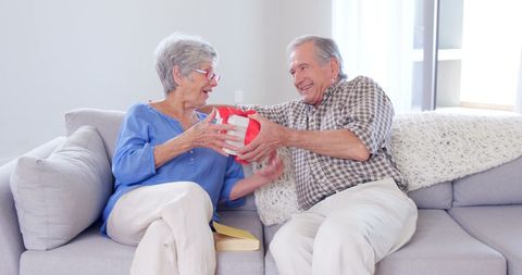 Elderly woman joyfully accepting gift from husband on sofa