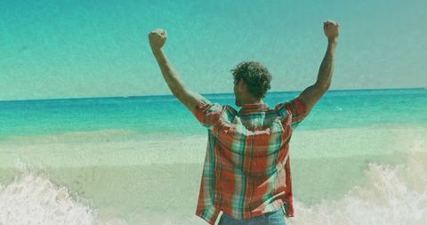 man standing on sandy beach with arms raised, expressing joy and freedom against shimmering turquoise ocean. ideal for concepts of success, vacation, enjoyment, or stress relief. suitable for travel blogs, motivational content, well-being promotions, or lifestyle advertising.