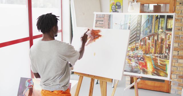 Man painting an urban landscape in bright, modern art studio. Canvas and other artworks surrounding him. Ideal for articles on creativity, urban art, artist lifestyle, or studio setup inspiration.