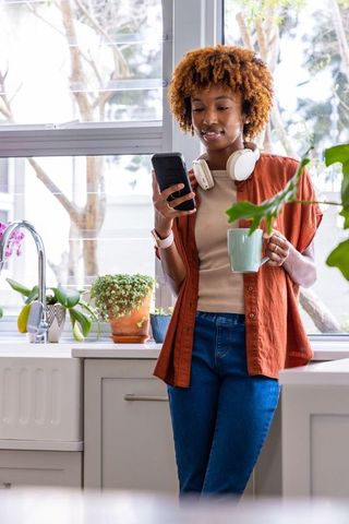 African american woman checking smartphone while holding mug in bright home kitchen