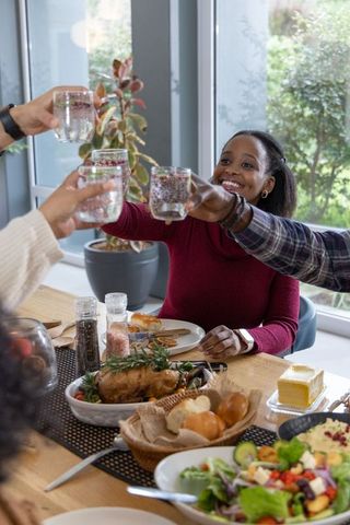 African american woman toasting friends at festive family dinner table with roast chicken