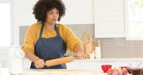 African american baker rolling dough in bright home kitchen wearing denim apron