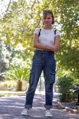 Young woman standing with arms crossed on sunlit park path in white tee, cargo jeans, sunglasses