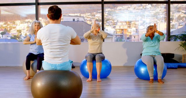 Senior individuals engage in a yoga session led by an instructor in a modern studio. They are seated on exercise balls, focusing on strength and balance training. The background features an urban cityscape through large windows. Ideal use for promotions of senior fitness programs, health and wellness campaigns, and exercise equipment marketing.