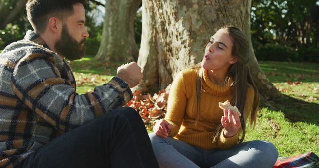 A young couple is sitting on the grass near a large oak tree, enjoying a picnic on a sunny autumn day. Both are dressed in casual clothing, and the woman is holding a sandwich while talking. Vibrant green and orange foliage of the season add to the pleasant ambiance, making it a great image for use in themes like leisure, nature, seasonal activities, healthy living, and romantic getaways.