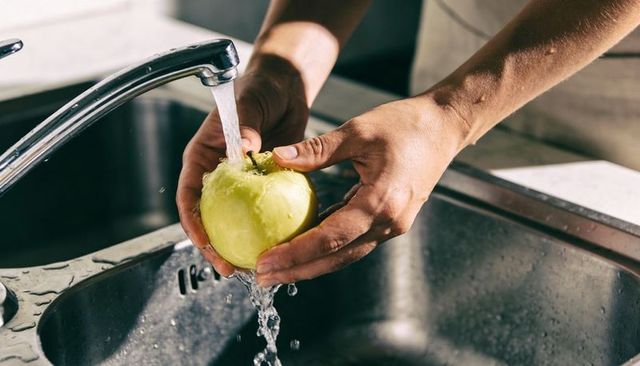 Rinsing green apple under running tap in kitchen sink with adult hands and apron