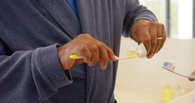 Close-up of man in bathrobe applying toothpaste to toothbrush, ideal for hygiene, personal care, morning routine, and wellness themed uses. Suitable for advertising oral hygiene products, promoting healthy lifestyle habits, or illustrating morning routines.