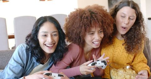 Laughing friends playing video games on couch with controllers and bowl of chips