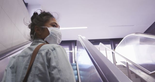 Young woman wearing face mask riding elevator in modern building, emphasizing health and safety measures during travel or commuting in urban environments. Suitable for themes related to public health, transportation, modern architecture, and everyday life during pandemics.