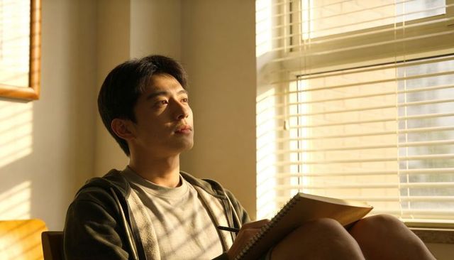 Young man journaling in sunlit room with blinds casting warm shadows, contemplating ideas