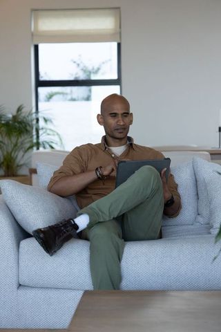 Middle-aged indian man sitting cross-legged on grey sofa using tablet in living room