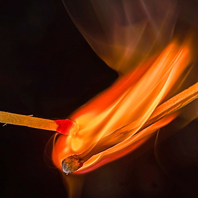 Intense close-up shot of two matchsticks where one is igniting the other. The contrast of the bright flame against the dark background captures attention, emphasizing the concepts of ignition, flame, and fire safety. This could be utilized in advertising related to fire safety, matches, campfires, or themes of ignition and energy. Ideal for educational materials, safety brochures, or dramatic commercial context.