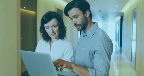 Colleagues Reviewing Laptop in Modern Office Hallway Showcasing Teamwork and Collaboration