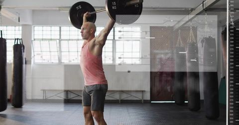 Male athlete pressing barbell overhead in industrial gym wearing coral tank and shorts