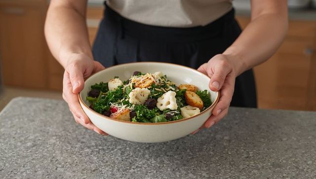 Woman presenting kale salad bowl with roasted cauliflower, croutons and cheese in kitchen