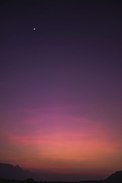 Captured during the early evening, the sky displays a palette of vibrant pink and purple hues while the crescent moon shines above darkened landscapes. Perfect for representations of tranquility, natural beauty, and the peaceful end of the day. Ideal for use in travel blogs, meditation app backgrounds, nature-themed websites, or inspirational quotes.