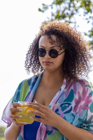 Sunlit african american woman wearing round sunglasses and colorful wrap holding citrus drink