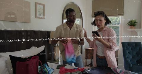 Young couple packing suitcase in cozy bedroom, coordinating travel plans via smartphone