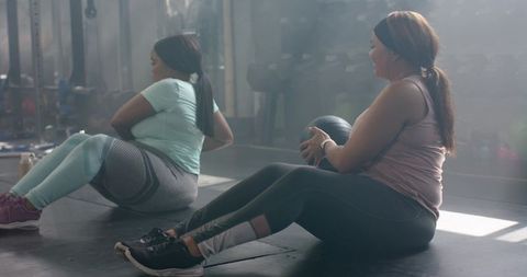 Diverse women doing seated medicine ball twists in gym training together in activewear