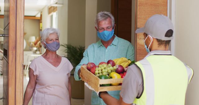 Elderly couple standing at their front door, wearing face masks, receiving a grocery delivery from a worker. Scene portrays safety measures during the coronavirus pandemic, emphasizing social distancing and use of protective gear. Useful for articles on home delivery, safety practices during pandemic, and services for senior citizens.