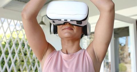Woman adjusting white vr headset on covered porch wearing sleeveless pink top
