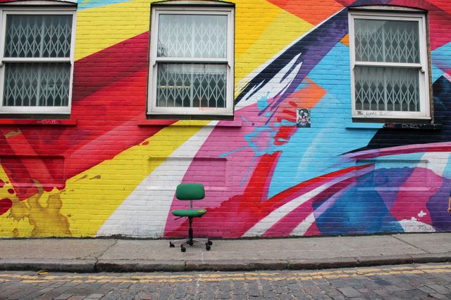 Vibrant street art featuring a colorful mural with a solitary green office chair under two windows on a city street. Great for designs about urban living, artistic inspiration, and modern art. Use for promoting events, city tours, or creative industries.