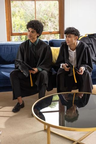 African american male graduates sitting on sofa holding mortarboards and tassels