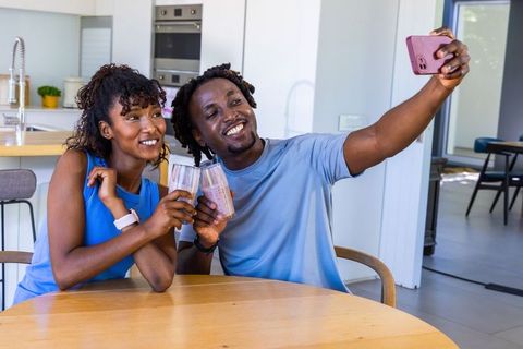 African american couple taking selfie while clinking smoothies in modern sunlit kitchen