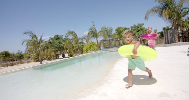 Children enjoying a sunny day by the swimming pool, wearing swimwear and holding colorful inflatable pool floats. Palm trees in the background enhance the tropical vacation atmosphere. Suitable for themes related to summer, vacations, outdoor activities, and childhood joy.