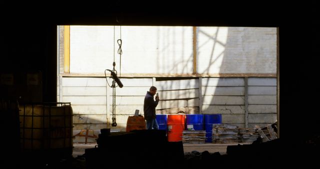 Worker stands in middle of industrial warehouse surrounded by barrels and equipment. Can be used to depict industrial work environments, safety procedures, and the daily life of industrial jobs in articles, websites, or presentations focusing on manufacturing, construction, or warehouse operations.