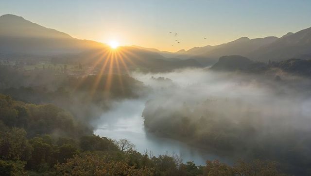 Misty river valley at sunrise casting sunbeams over forested mountains and hills