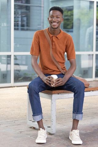Smiling african american man sitting on bench holding takeaway coffee by glass facade
